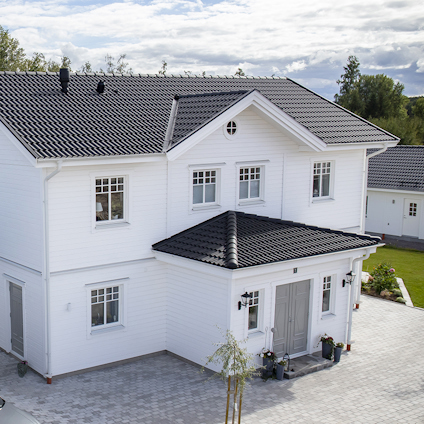 Tvåvånings hus med vita träfasader, svarta takpannor och en stensatt uppfart. Omgivande grönska och molnig himmel.
